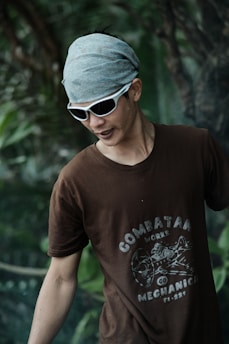 A person is wearing a brown t-shirt with a graphic design and text on it, sunglasses, and a gray headscarf. The background is filled with green foliage, suggesting a natural or outdoor setting.