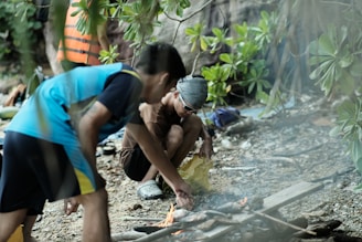 Several people are outdoors near a makeshift fire. One person is adjusting something on the ground near the fire, possibly preparing food or maintaining the fire. Another person nearby is wearing a life jacket. The area is surrounded by dense green foliage and scattered rocks.