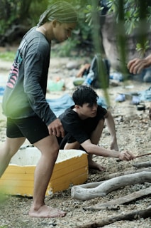 Two people are on a pebbled shoreline, engaging in an activity possibly related to outdoor exploration or survival. One person crouches while reaching out towards driftwood, and the other stands nearby. There are various outdoor items scattered around, including a yellow and white piece of material that could be a section of a boat or container.