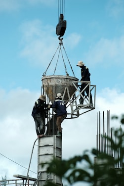 Three construction workers are balancing on scaffolding high above the ground, handling a large cement bucket suspended by a crane. The sky is clear and blue, providing a backdrop to the scene of industrial work.