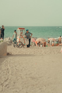 On a sandy beach, a vendor with a blue cap stands next to a colorful popcorn cart, while people enjoy the beach under umbrellas near the ocean. Several beachgoers are visible, some standing and others sitting, enjoying the relaxed atmosphere.