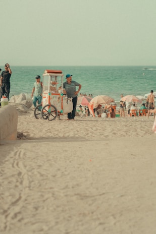 On a sandy beach, a vendor with a blue cap stands next to a colorful popcorn cart, while people enjoy the beach under umbrellas near the ocean. Several beachgoers are visible, some standing and others sitting, enjoying the relaxed atmosphere.