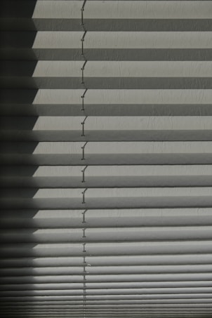 Horizontal window blinds casting layered shadows with alternating light and dark sections, forming a pattern on the surface.
