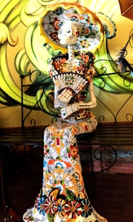 A decorative skeleton figure is elegantly dressed in a richly patterned, colorful gown and wide-brimmed hat. The skeleton is seated, holding a fan, and features intricate floral designs. The background showcases swirling green patterns.