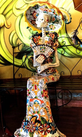 A decorative skeleton figure is elegantly dressed in a richly patterned, colorful gown and wide-brimmed hat. The skeleton is seated, holding a fan, and features intricate floral designs. The background showcases swirling green patterns.