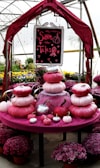 A decorative setup featuring stacked pumpkins in shades of pink, placed on a round table covered with pink fabric. Above them is a sign reading 'Save the Tatas' with heart motifs and a pink ribbon. The background shows a greenhouse environment with potted flowers in vibrant colors, enhancing the lively atmosphere.