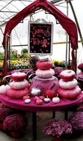 A decorative setup featuring stacked pumpkins in shades of pink, placed on a round table covered with pink fabric. Above them is a sign reading 'Save the Tatas' with heart motifs and a pink ribbon. The background shows a greenhouse environment with potted flowers in vibrant colors, enhancing the lively atmosphere.