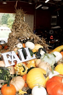 A rustic arrangement of pumpkins and gourds in various sizes and colors is accompanied by a decorative metallic sign spelling 'FALL.' The display is topped with a stack of dried corn stalks, creating a warm and festive autumn atmosphere inside a wooden barn.
