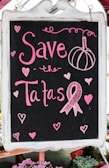 A blackboard with pink chalk writing features the phrase 'Save the Tatas' accompanied by decorative elements like hearts and a ribbon symbol often associated with breast cancer awareness. The board is framed in white and surrounded by a few blurred flowers at the bottom.