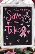 A blackboard with pink chalk writing features the phrase 'Save the Tatas' accompanied by decorative elements like hearts and a ribbon symbol often associated with breast cancer awareness. The board is framed in white and surrounded by a few blurred flowers at the bottom.