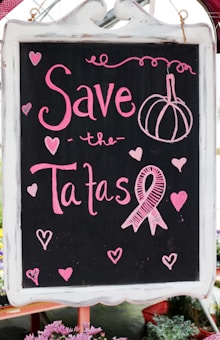 A blackboard with pink chalk writing features the phrase 'Save the Tatas' accompanied by decorative elements like hearts and a ribbon symbol often associated with breast cancer awareness. The board is framed in white and surrounded by a few blurred flowers at the bottom.