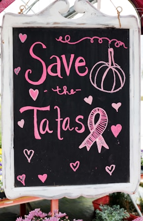 A blackboard with pink chalk writing features the phrase 'Save the Tatas' accompanied by decorative elements like hearts and a ribbon symbol often associated with breast cancer awareness. The board is framed in white and surrounded by a few blurred flowers at the bottom.