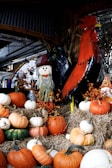 A display of autumn harvest decorations featuring various pumpkins in shades of orange, white, and green arranged on straw bales. There's a decorative scarecrow with a hat and a large, colorful metal rooster. Small orange berries add to the seasonal theme.
