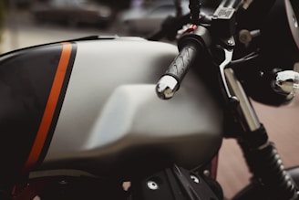 A close-up view of a motorcycle with a sleek, metallic fuel tank featuring a bold orange stripe. The handlebars and throttle grip appear prominently, highlighting the bike's polished chrome and black details. The blurred background suggests an outdoor setting.