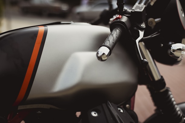 A close-up view of a motorcycle with a sleek, metallic fuel tank featuring a bold orange stripe. The handlebars and throttle grip appear prominently, highlighting the bike's polished chrome and black details. The blurred background suggests an outdoor setting.