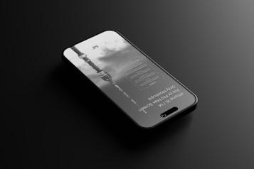 A sleek smartphone lying flat on a dark, reflective surface. The screen displays text and a grayscale image of a tower or building against a cloudy sky. The design of the phone appears modern with a thin profile and a visible front camera cutout.