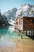 A wooden cabin built on stilts extends over clear turquoise water with majestic mountains in the background. Small boats are visible on the tranquil lake, while the sunlight casts a warm glow on the scene.