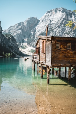 A wooden cabin built on stilts extends over clear turquoise water with majestic mountains in the background. Small boats are visible on the tranquil lake, while the sunlight casts a warm glow on the scene.