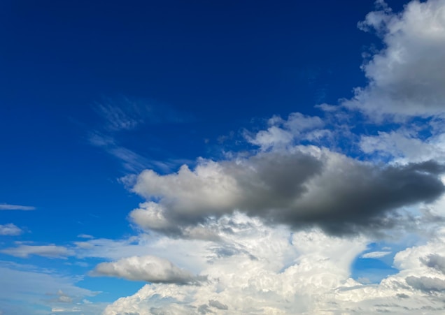 A clear sky with varying shades of blue, featuring large, fluffy white clouds and some darker, more ominous clouds. The bright blue provides a backdrop that highlights the intricate shapes and textures of the clouds, suggesting a dynamic and ever-changing weather pattern.