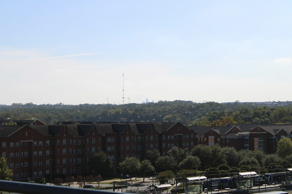 The image features a large multi-story brick building in the foreground with a pitched roof, surrounded by lush green trees and landscaping. In the background, there is a densely wooded area that extends towards the horizon. Additionally, two tall communication towers are visible in the distance against a clear blue sky. Below the building, there's a street or tramway station with several vehicles or structures lining up.