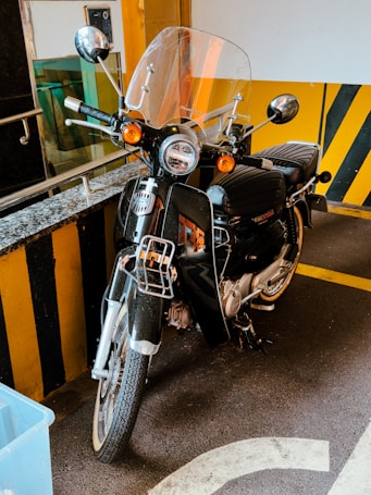 A parked motorcycle is positioned against a yellow and black striped barrier. The motorcycle, predominantly black with chrome details, is equipped with a windshield, side mirrors, and orange signal lights. Behind the motorcycle, a granite countertop and handrail are visible. The floor has yellow parking lines and white markings.
