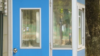 A small blue and white booth with large windows is positioned outdoors. The background features soft focus greenery and blurred figures, suggesting a park or open area setting. The booth is partially obscured by a tree trunk on the right side.