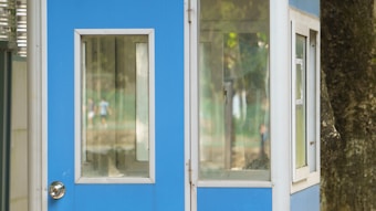 A small blue and white booth with large windows is positioned outdoors. The background features soft focus greenery and blurred figures, suggesting a park or open area setting. The booth is partially obscured by a tree trunk on the right side.