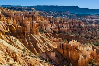 A vast landscape featuring dramatic geological formations known as hoodoos, with towering rock spires in varying shades of orange and brown. The scene includes a backdrop of distant mountains under a clear blue sky, with scattered green vegetation adding contrast to the arid terrain.