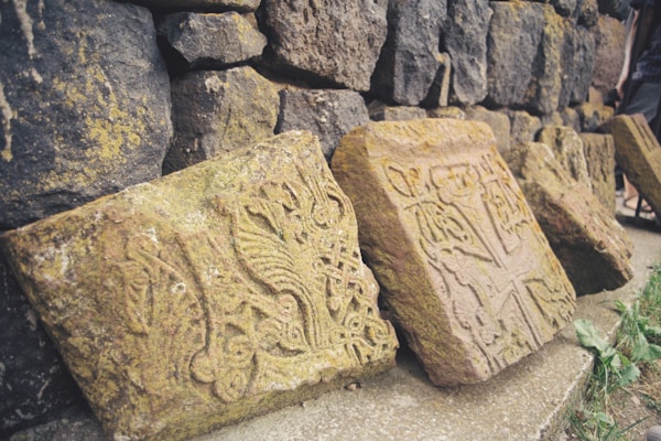 Stone blocks with intricate carvings, leaning against a weathered stone wall. The carvings appear to be historical or decorative motifs. Some grass and small plants can be seen at the base, suggesting an outdoor setting.