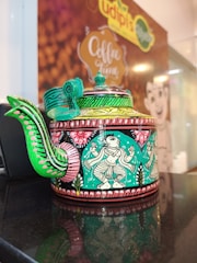 A decorative teapot featuring intricate, colorful artwork with a cultural motif. The design includes traditional patterns and figures with vibrant colors like green and pink covering the surface. It is placed on a reflective surface, enhancing its ornate details.