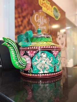 A decorative teapot featuring intricate, colorful artwork with a cultural motif. The design includes traditional patterns and figures with vibrant colors like green and pink covering the surface. It is placed on a reflective surface, enhancing its ornate details.