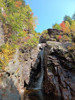 A scenic waterfall cascades down a rocky cliff surrounded by vibrant autumn foliage with shades of red, orange, and green. The bright blue sky serves as a backdrop to the lush natural setting.