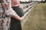 A pregnant person stands next to a wooden fence in a grassy area. The person is wearing a floral-patterned garment with lace details over a pink top and dark green skirt. They are gently holding their belly with both hands.