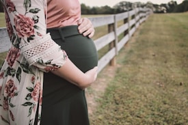 A pregnant person stands next to a wooden fence in a grassy area. The person is wearing a floral-patterned garment with lace details over a pink top and dark green skirt. They are gently holding their belly with both hands.