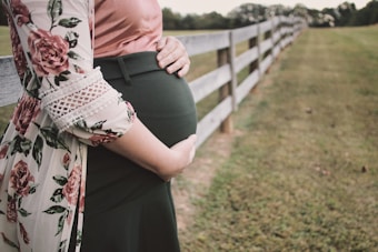 A pregnant person stands next to a wooden fence in a grassy area. The person is wearing a floral-patterned garment with lace details over a pink top and dark green skirt. They are gently holding their belly with both hands.