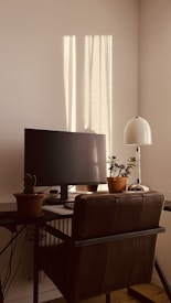 A cozy workspace featuring a computer monitor on a desk, accompanied by potted plants. The area is bathed in warm sunlight streaming through a window, casting shadows on the wall. A modern desk lamp adds to the setting, and a dark leather chair is positioned in front of the desk.