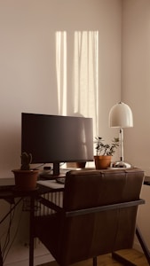 A cozy workspace featuring a computer monitor on a desk, accompanied by potted plants. The area is bathed in warm sunlight streaming through a window, casting shadows on the wall. A modern desk lamp adds to the setting, and a dark leather chair is positioned in front of the desk.