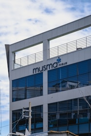A modern office building with a sign saying 'musma' and additional Korean text. The structure features reflective glass windows, a rooftop railing, and a light gray facade. A power line with a utility pole is visible in the foreground.
