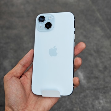 A hand is holding a white smartphone with dual cameras and an Apple logo on the back.
