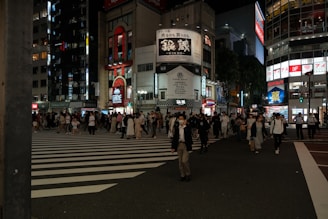 A bustling urban street scene at night with numerous pedestrians walking across a wide zebra crossing. The backdrop features tall buildings with illuminated signage and billboards displaying various advertisements. The scene is lively, with a mix of people in casual and business attire, indicating a busy district.