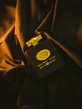 A black and gold cologne bottle rests on dark, crumpled fabric. The bottle has elegant golden details and lettering that enhance its luxurious appearance. The lighting casts rich shadows and highlights, contributing to a sophisticated ambiance.