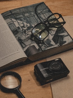 A vintage-style composition featuring an open book with a detailed illustration, a pair of glasses resting on the pages, a magnifying glass placed on a wooden table, and a Fujifilm camera beside it. The scene evokes a sense of exploration and nostalgia.