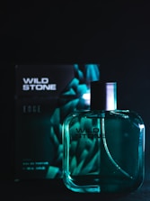 A bottle of Wild Stone perfume is positioned in front of its packaging. The bottle is translucent with a dark spray nozzle, and the packaging behind features a geometric design. The setting is dark, with teal and black tones dominating the scene.