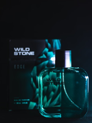 A bottle of Wild Stone perfume is positioned in front of its packaging. The bottle is translucent with a dark spray nozzle, and the packaging behind features a geometric design. The setting is dark, with teal and black tones dominating the scene.