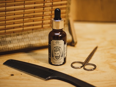 A bottle of beard oil with a dropper cap is placed on a wooden surface. To the right, a pair of metal scissors rests nearby. A black comb lies flat on the left, and behind the items, there is a woven bamboo-like mat.