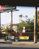A street scene with a bus stop featuring two advertisements, one for a service and another for a show or movie. A person is seated on the bench at the bus stop. In the background, there are palm trees, bushes with red flowers, and residential buildings. The lighting suggests it is either early morning or late afternoon.