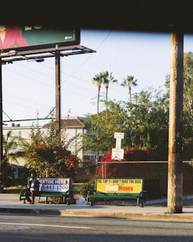 A street scene with a bus stop featuring two advertisements, one for a service and another for a show or movie. A person is seated on the bench at the bus stop. In the background, there are palm trees, bushes with red flowers, and residential buildings. The lighting suggests it is either early morning or late afternoon.