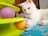 A white cat is playing with a toy that has colorful balls inside. The background includes a textured surface and some indoor elements.