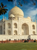 The image features the iconic marble structure of the Taj Mahal, showcasing its large dome and detailed architectural carvings. The foreground includes a lush green lawn with people walking and admiring the monument. The building's pristine white marble contrasts with the vibrant blue sky and the greenery surrounding it.