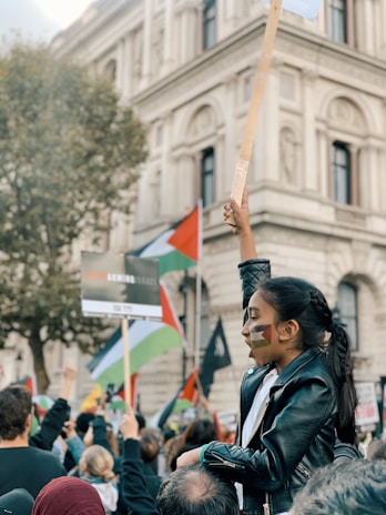A group of people is gathered in front of a historical building, participating in a protest. The crowd is holding signs and flags, including noticeable Palestinian flags. A young girl with painted face marks is sitting on someone's shoulders, raising a sign with determination.